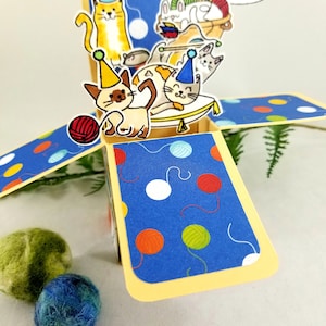 May include: A pop-up birthday card featuring cartoon cats wearing party hats. The card has a blue background with colorful yarn ball designs. The text "Happy Purr-thday" is in a speech bubble. Two felt balls are in the foreground.