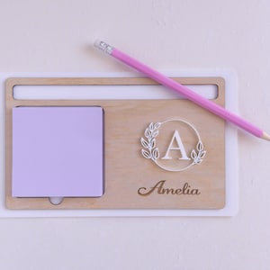 May include: A wooden desk organiser with a light wood finish. It has a space for a notepad, a pencil holder, and a personalised nameplate with the name "Amelia" and a decorative letter "A" with leaves.