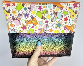 May include: A rectangular cosmetic bag with a colorful roller skate and star pattern on a white background. The bottom half is covered in rainbow glitter. The bag has an orange zipper and is held by a hand.