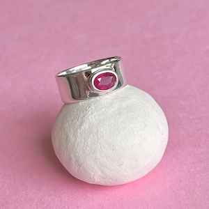 Może przedstawiać: Srebrny pierścionek z czerwonym, owalnym kamieniem osadzonym w środku. Pierścionek jest gruby i ma zaokrągloną obręcz.