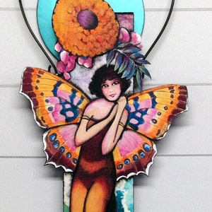 May include: A colorful wooden wall hanging featuring a whimsical fairy with butterfly wings, surrounded by flowers and a blue circle.