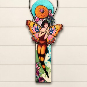 May include: A vertical wall hanging featuring a central figure with large, colorful butterfly wings in shades of orange, pink, and yellow. The figure is positioned against a rectangular panel with a teal circular accent, orange flower, and floral details.