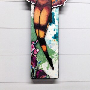 May include: A rectangular wall art piece featuring a stylized illustration of a moth with orange and black wings. The lower section displays vibrant purple and pink flowers against a textured white, green, and blue watercolor-style background.