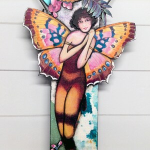 May include: A decorative wall hanging featuring a central figure with dark hair and large, vibrant butterfly wings in shades of orange, pink, and blue. The figure wears a dark, form-fitting outfit against a rectangular background with teal and white patterns.