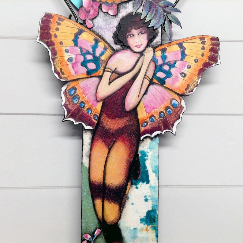 May include: A decorative wall hanging featuring a central figure with dark hair and large, vibrant butterfly wings in shades of orange, pink, and blue. The figure wears a dark, form-fitting outfit against a rectangular background with teal and white patterns.