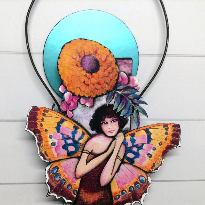May include: A decorative wall hanging featuring a central illustration of a person with dark hair and butterfly wings in shades of orange, pink, and blue. Above the figure is a large orange flower and a bright turquoise circular background element.
