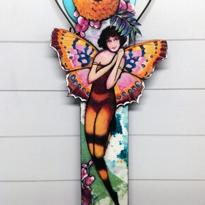 May include: A decorative wall hanging featuring a central figure with dark hair and large, vibrant orange and pink butterfly wings. The figure is positioned against a rectangular panel with a blue circular accent, floral details, and green foliage.