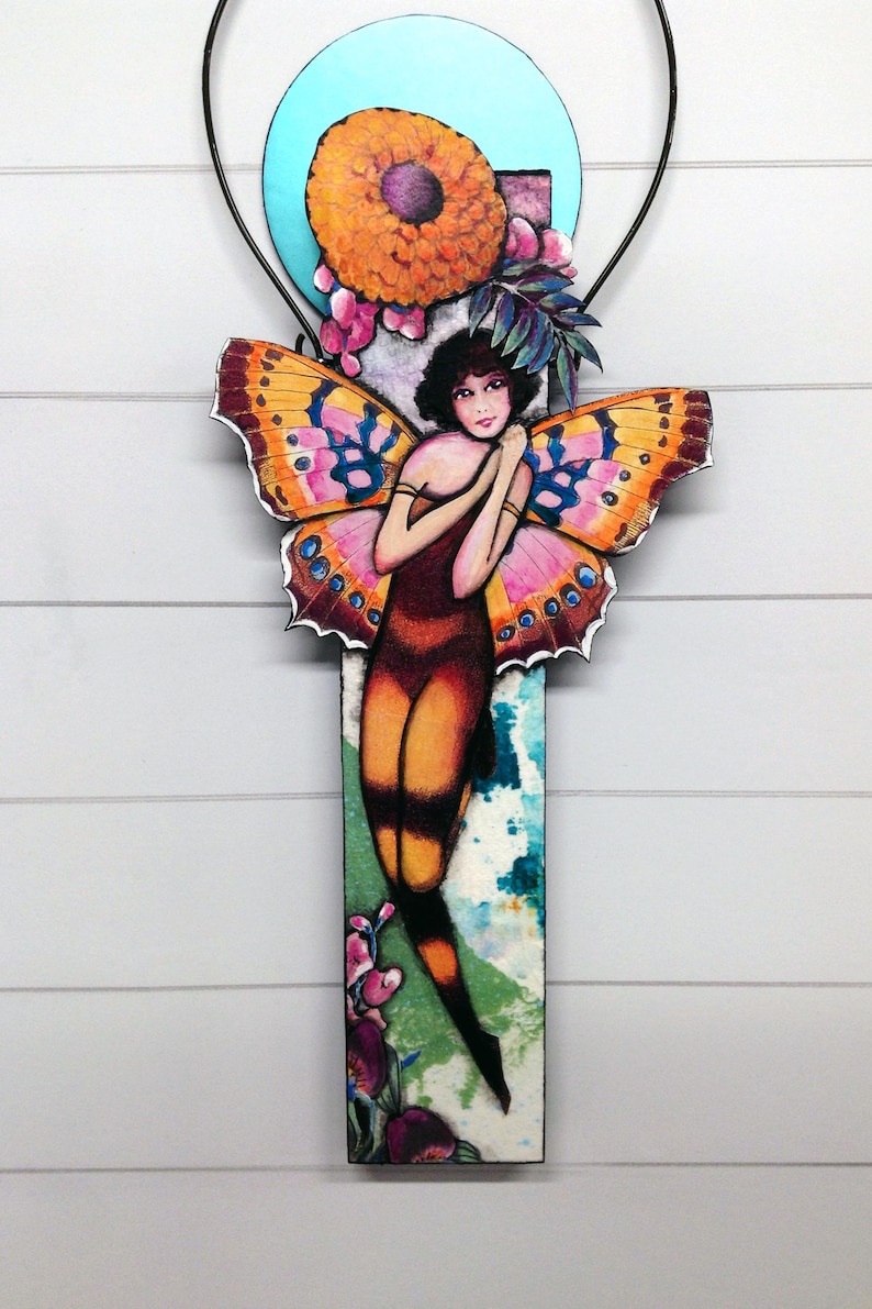 May include: A decorative wall hanging featuring a central figure with dark hair and large, vibrant orange and pink butterfly wings. The figure is positioned against a rectangular panel with a blue circular accent, floral details, and green foliage.
