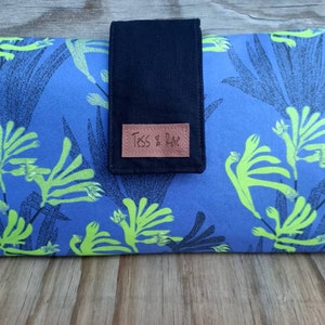 May include: A blue clutch purse with a black flap and a brown leather tag that says "Tess & Rae". The purse has a floral pattern with green and white flowers.