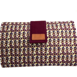 May include: A burgundy clutch with a geometric pattern in shades of gold, white, and purple. The clutch has a burgundy flap with a brown leather label.