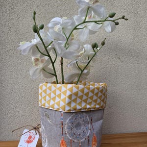 May include: A white orchid plant in a grey and yellow fabric planter with a dreamcatcher design. The planter has a yellow and white geometric pattern on the top edge.