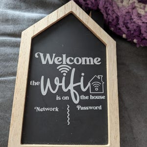 May include: A wooden sign shaped like a house with a chalkboard insert. The sign says "Welcome the wifi is on the house" with a wifi symbol and a house icon. The words "Network" and "Password" are written below with blank lines for the user to fill in.