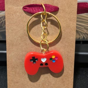 May include: Red plastic video game controller keychain with a gold chain and ring. The controller has a black plus sign, a white heart, and colorful buttons.