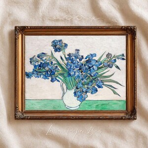 May include: A framed art print featuring a bouquet of vibrant blue irises with long green leaves arranged in a white pitcher. The pitcher sits on a flat green surface against a textured off-white background, all enclosed in an ornate gold frame.