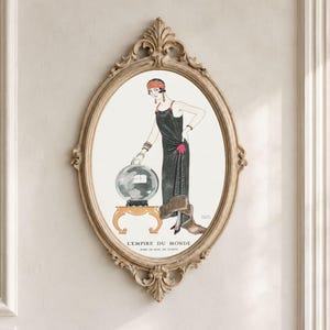Może przedstawiać: Owalny obraz ścienny przedstawiający ilustrację postaci w czarnej sukni z futrzaną etolą, stojącej obok kryształowej kuli na małym stoliku. Na dole znajduje się napis L'EMPIRE DU MONDE oraz ROSE DU SOIR, DE WORTH.
