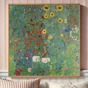 Puede incluir: Una impresi&oacute;n enmarcada de una escena de jard&iacute;n vibrante, con girasoles, flores rojas, blancas y azules, y exuberante follaje verde. La obra de arte est&aacute; en un marco cuadrado, dorado, y se exhibe contra una pared a rayas rosas y blancas.