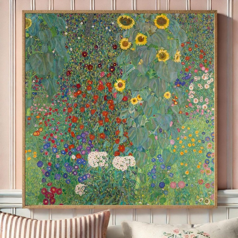 Puede incluir: Una impresi&oacute;n enmarcada de una escena de jard&iacute;n vibrante, con girasoles, flores rojas, blancas y azules, y exuberante follaje verde. La obra de arte est&aacute; en un marco cuadrado, dorado, y se exhibe contra una pared a rayas rosas y blancas.