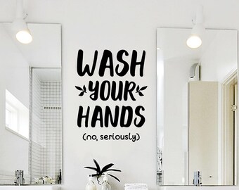 以下が含まれることがあります： WASH YOUR HANDSという文字と、YOURという単語の両側に小さな葉のアイコン、その下に小さなフォントで(no, seriously)と書かれた黒いビニール製のウォールステッカー。2枚の長方形の鏡の間の白い壁に貼られている。