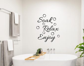 May include: A black wall decal featuring the words Soak, Relax, and Enjoy in a cursive script font, interspersed with various circular bubble shapes. The decal is applied to a plain white wall above a white bathtub with a wooden tray.
