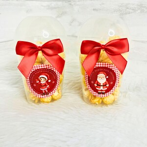 May include: Two clear plastic cups filled with popcorn, each topped with a red satin bow. The left cup features a circular label with an elf illustration and the text "MAGICAL NORTH POLE DELIVERY". The right cup has a Santa Claus illustration.