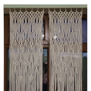 May include: Two white macrame curtains hanging in a window. The curtains are made of a thick, knotted rope and have a geometric pattern. The curtains are hanging in a window with a wooden frame.