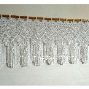 May include: White macrame wall hanging with a geometric pattern and fringe. The hanging is attached to a wooden dowel rod.