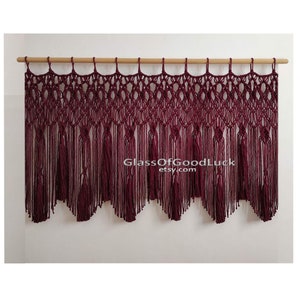 Pode incluir: Uma tapeçaria de macramé de cor bordô com uma haste de madeira na parte superior. A tapeçaria apresenta um design de franjas com borlas. O texto "GlassOfGoodLuck etsy.com" é visível no centro da tapeçaria.