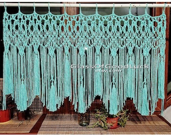 May include: A turquoise macrame curtain with tassels hanging from a white rod. The curtain is made of knotted string and has a diamond pattern. The curtain is hanging in front of a window with a wooden frame.