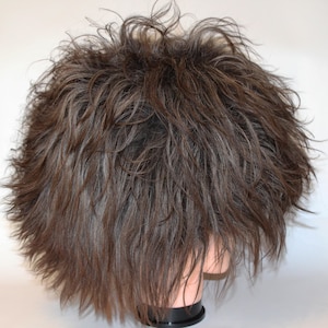 May include: A brown wig with a short, layered style. The wig has a textured, tousled appearance with strands of grey hair mixed in. The wig is on a mannequin head with a black base, suitable for styling and display.