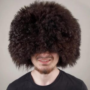 Può includere: Una persona indossa un grande cappello marrone scuro e soffice che copre gli occhi. La persona indossa una maglietta nera e ha la barba. Lo sfondo è di colore chiaro.