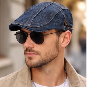 Puede incluir: Gorra de estilo newsboy de mezclilla azul con visera curva. La gorra tiene costuras visibles y un pequeño detalle de correa en el lateral. La persona de la imagen lleva gafas de sol de aviador negras y una chaqueta color beige.
