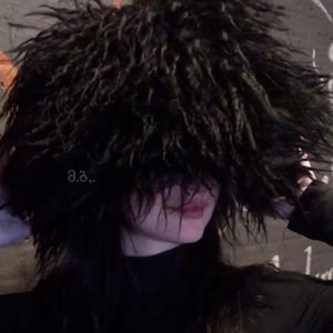 Può includere: Una persona indossa un cappello nero e soffice che le copre gli occhi. Il cappello è molto spesso e soffice. La persona indossa una maglietta nera a collo alto.