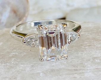 May include: A silver engagement ring with a large emerald cut diamond and two smaller pear-shaped diamonds on the sides.