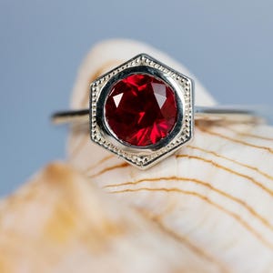 May include: A silver ring with a hexagonal setting featuring a vibrant red gemstone. The ring is set against a blurred background, highlighting the jewelry's design and color. The gemstone is a deep red color.