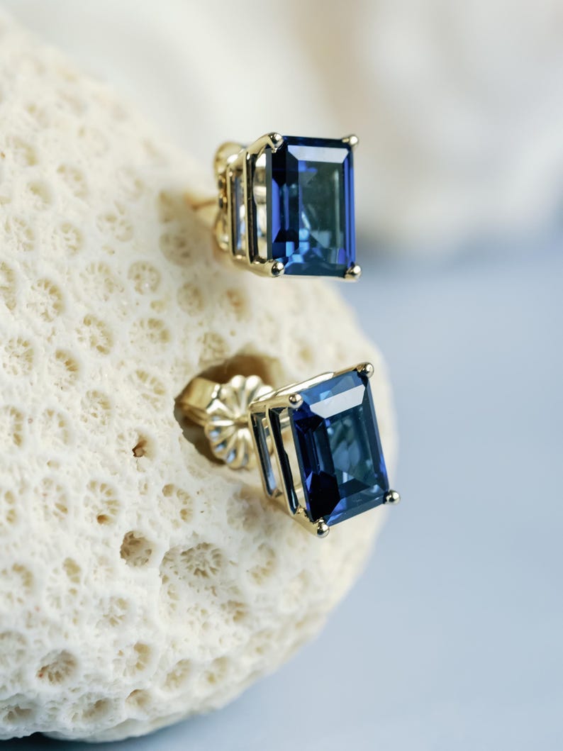 Puede incluir: Un par de pendientes de oro con piedras preciosas azules. Las piedras preciosas son de corte esmeralda y tienen un color azul oscuro.