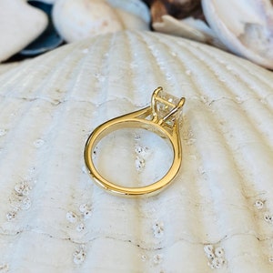 May include: A gold engagement ring with a square-shaped diamond set in a prong setting. The ring is on a white seashell background.