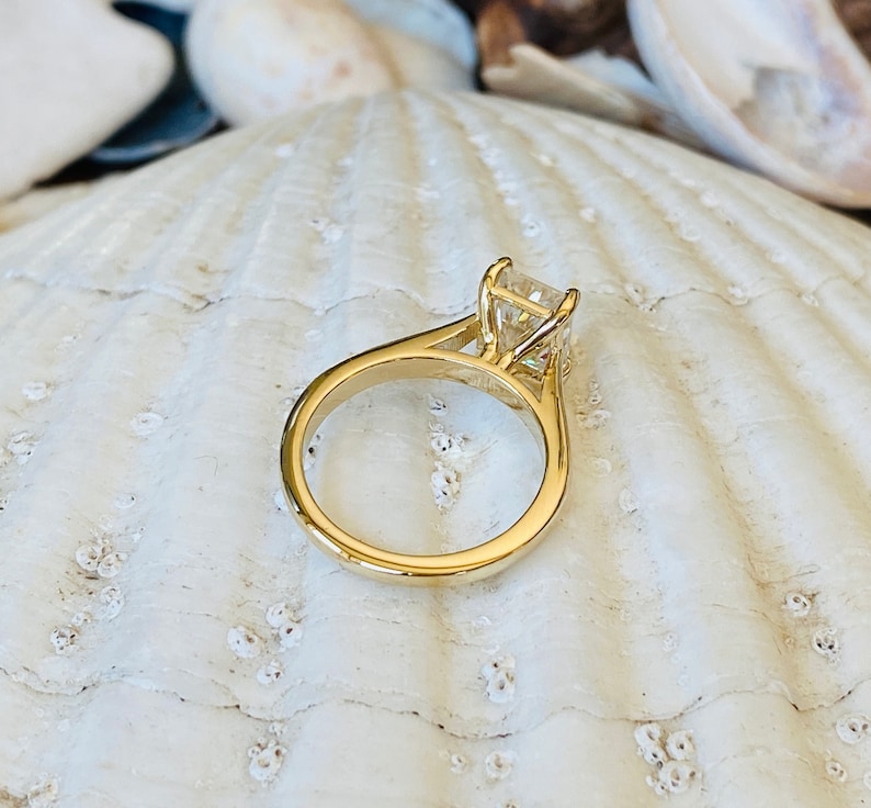 May include: A gold engagement ring with a square-shaped diamond set in a prong setting. The ring is on a white seashell background.