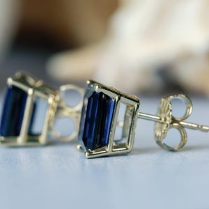 Puede incluir: Un par de pendientes de oro con piedras preciosas azules. Las piedras preciosas son de corte esmeralda y engastadas en un marco cuadrado.