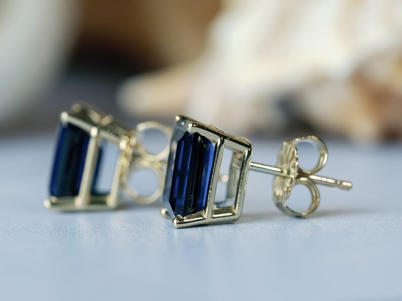 Puede incluir: Un par de pendientes de oro con piedras preciosas azules. Las piedras preciosas son de corte esmeralda y engastadas en un marco cuadrado.