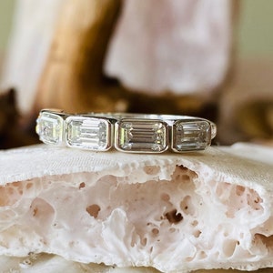 May include: A silver band ring with five emerald-cut diamonds. The ring is set on a white, textured surface.