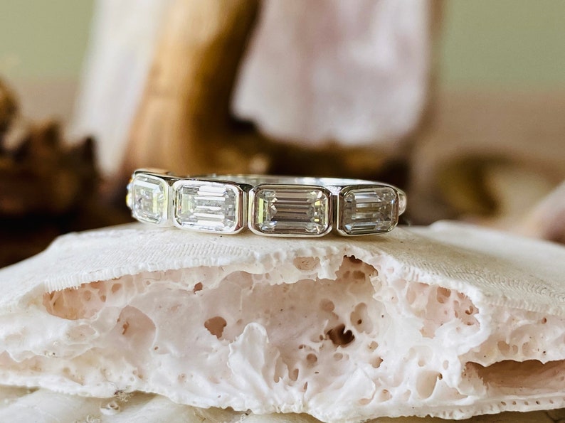 May include: A silver band ring with five emerald-cut diamonds. The ring is set on a white, textured surface.