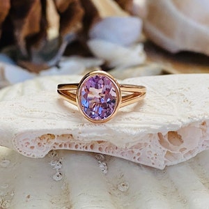 Pode incluir: Um delicado anel de ouro rosa com uma gema oval de cor púrpura clara. O anel está colocado sobre um fundo de concha branca.