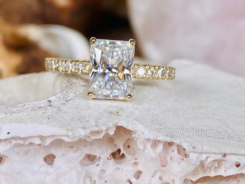 Pode incluir: Um anel de noivado de ouro com um grande diamante transparente de forma retangular. O anel tem uma banda de pequenos diamantes.