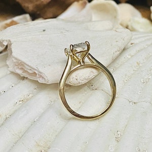 May include: A gold engagement ring with a square-shaped diamond set in a prong setting. The ring is on a white seashell background.
