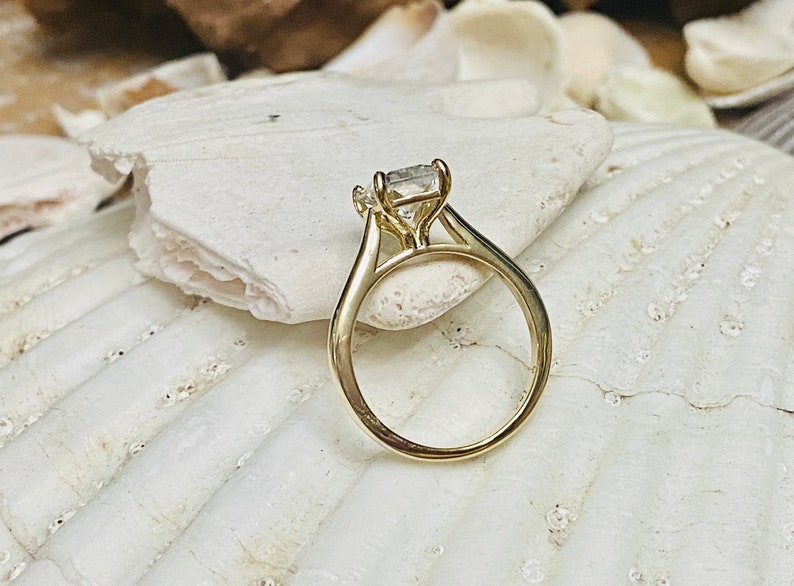 May include: A gold engagement ring with a square-shaped diamond set in a prong setting. The ring is on a white seashell background.