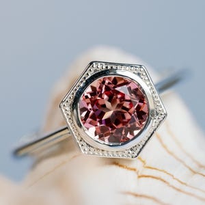 May include: A silver ring with a hexagonal setting. The ring features a round, faceted, pink gemstone. The setting is detailed with small, decorative elements. The ring is displayed on a light-colored, textured surface.