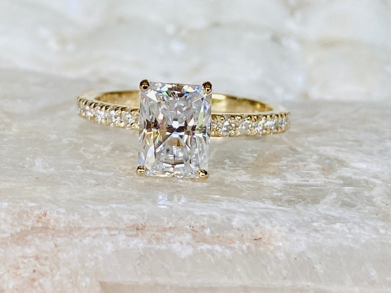 Pode incluir: Um anel de noivado de ouro com uma grande pedra central de diamante retangular e transparente, e uma banda de diamantes menores.