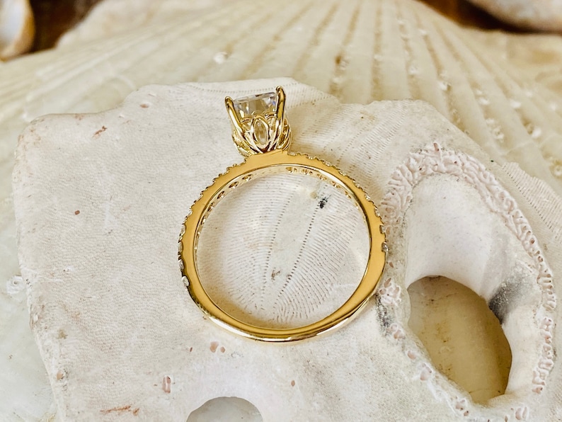 Pode incluir: Um anel de ouro com uma gema transparente engastada em garras. A banda &eacute; adornada com pequenas pedras brilhantes.