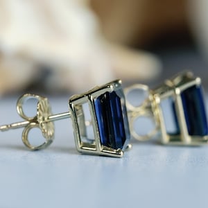 Puede incluir: Un par de pendientes de oro con piedras preciosas de color azul oscuro talla esmeralda. Los pendientes tienen un cierre de poste.