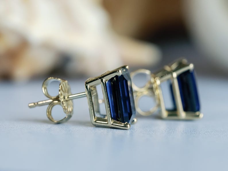 Puede incluir: Un par de pendientes de oro con piedras preciosas de color azul oscuro talla esmeralda. Los pendientes tienen un cierre de poste.
